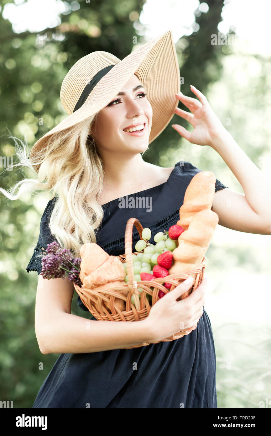 Smiling blonde femme 24-29 ans wearing blue robe et chapeau de paille holding corbeille de fruits, du pain frais à l'extérieur. À l'avant. 20s. Banque D'Images