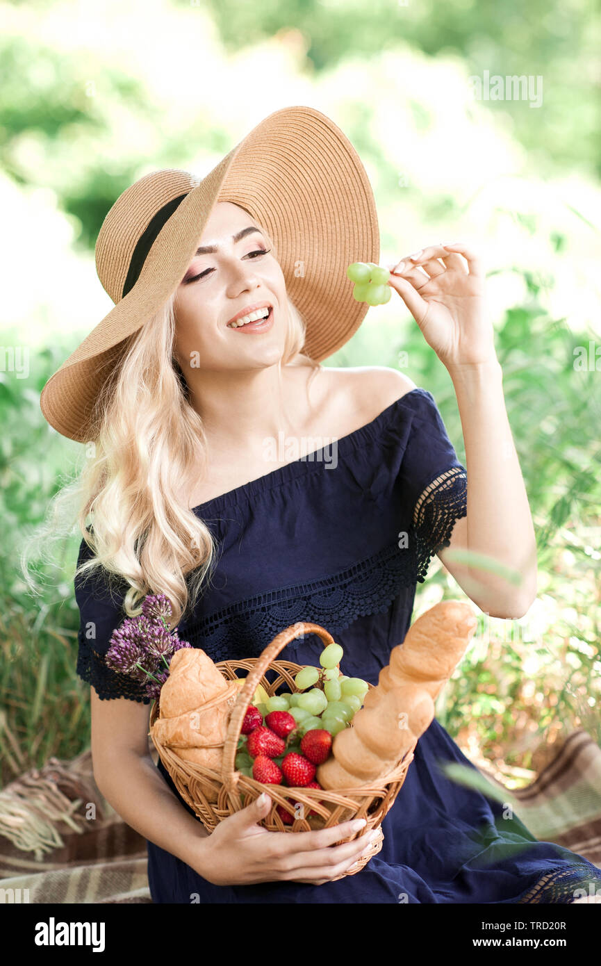 Smiling girl 24-29 ans avoir pique-nique au parc. Holding panier en osier avec des fruits et du pain. In et élégante robe bleue à l'extérieur. Somme Banque D'Images