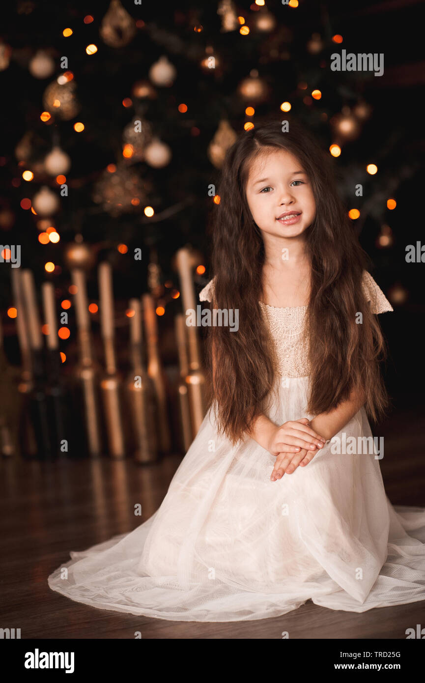 Mignon bebe fille 4 5 ans portant robe elegante assise sous les lumieres de Noel sur l arbre de Noel. En regardant la camera. Saison de vacances Photo Stock Alamy