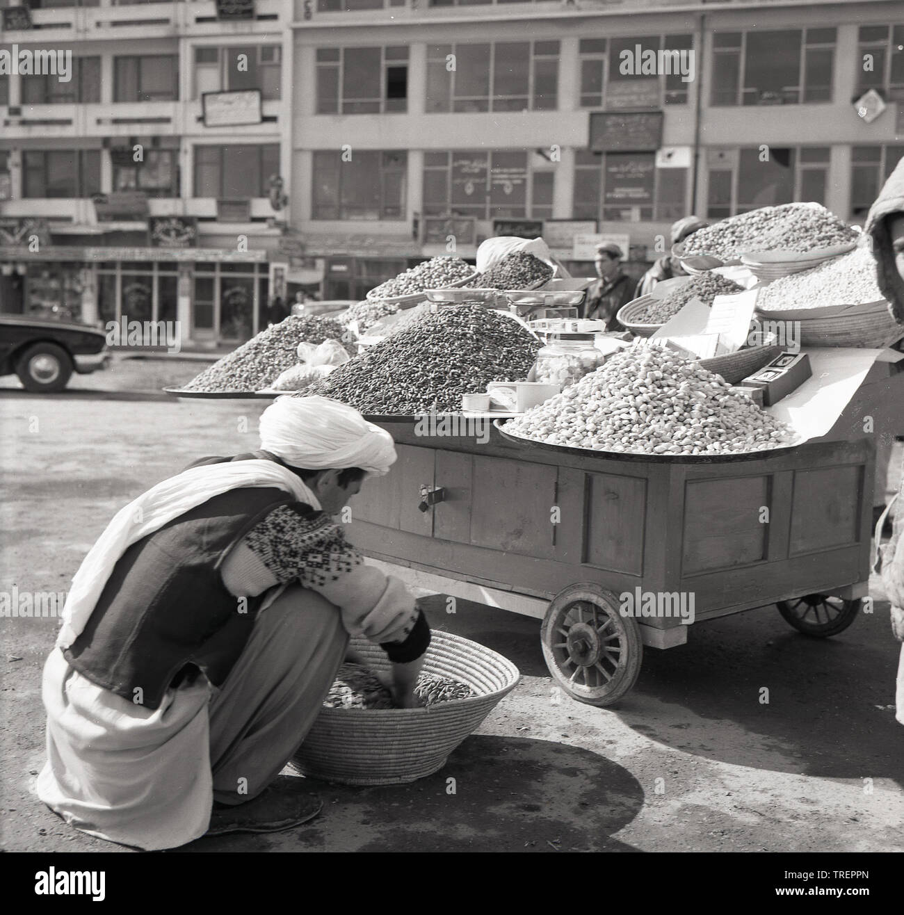 1950s, historique, à l'extérieur d'une rue à Kabal, en Afghanistan, un négociant afghan sur le terrain triant un panier de ses produits, différents types de noix, par sa stalle, un chariot mobile. Banque D'Images