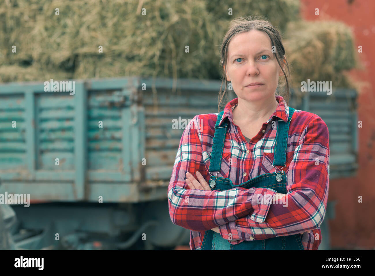 Female farmer posing in front of charrette à foin. Portrait de femme  travailleur agricole en chemise à carreaux et salopette par le tracteur  remorque remplie de beaucoup de produits laitiers Photo Stock -