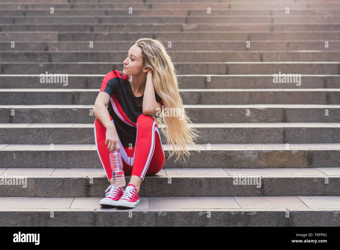 Jeune femme dans des vêtements tout assis sur des escaliers avec une bouteille d'eau. Elle porte un pantalon rouge et d'exercice chaussures rouges. Banque D'Images