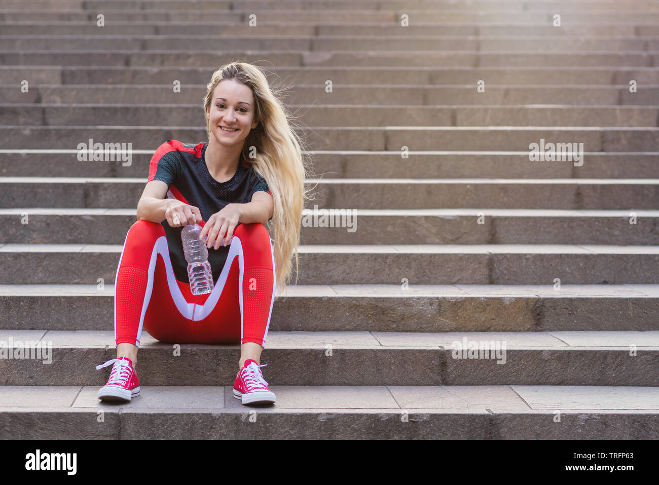 Jeune femme dans des vêtements tout assis sur des escaliers avec une bouteille d'eau. Elle porte un pantalon rouge et d'exercice chaussures rouges. Banque D'Images