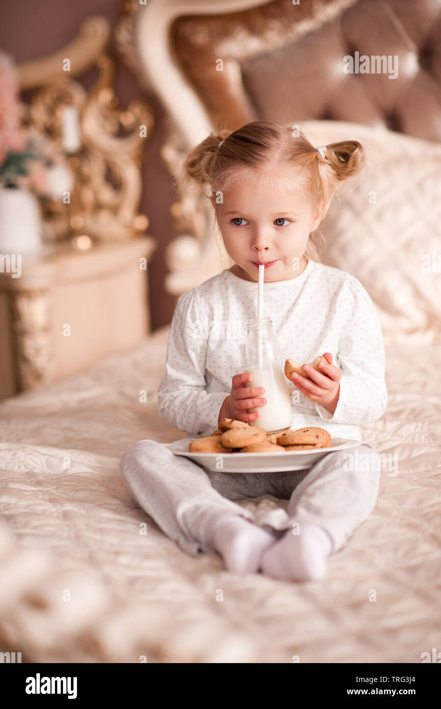Mignon bébé fille 3-4 ans boire du lait et manger des cookies dans le lit. Bonjour. L'heure du petit-déjeuner. Banque D'Images