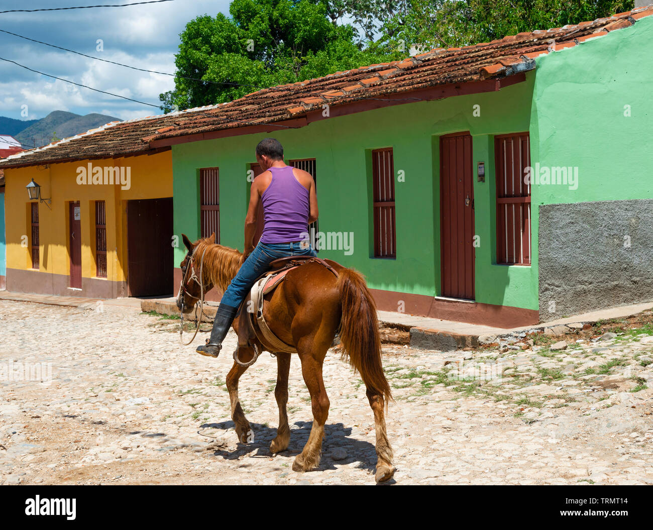 Cowboy chevauche son cheval le long de la rue pavée bordée de maisons colorées avec des montagnes Escambray en arrière-plan - Trinidad, Cuba, Caraïbes Banque D'Images