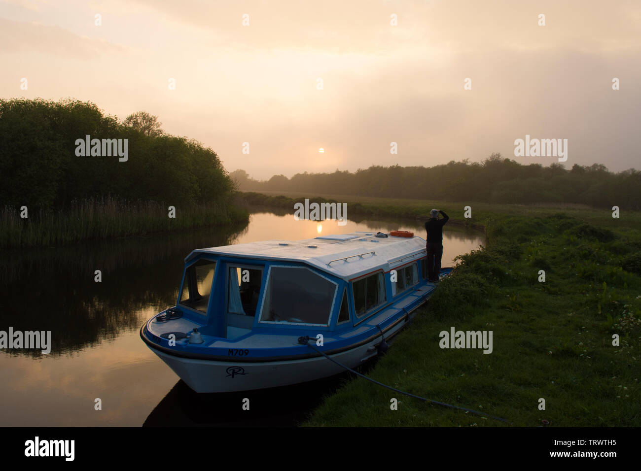 Maison de vacances sur l'homme sur le pont du bateau de plaisance de regarder un coucher de soleil sur la rivière ant, les Norfolk Broads, UK, mai. Banque D'Images
