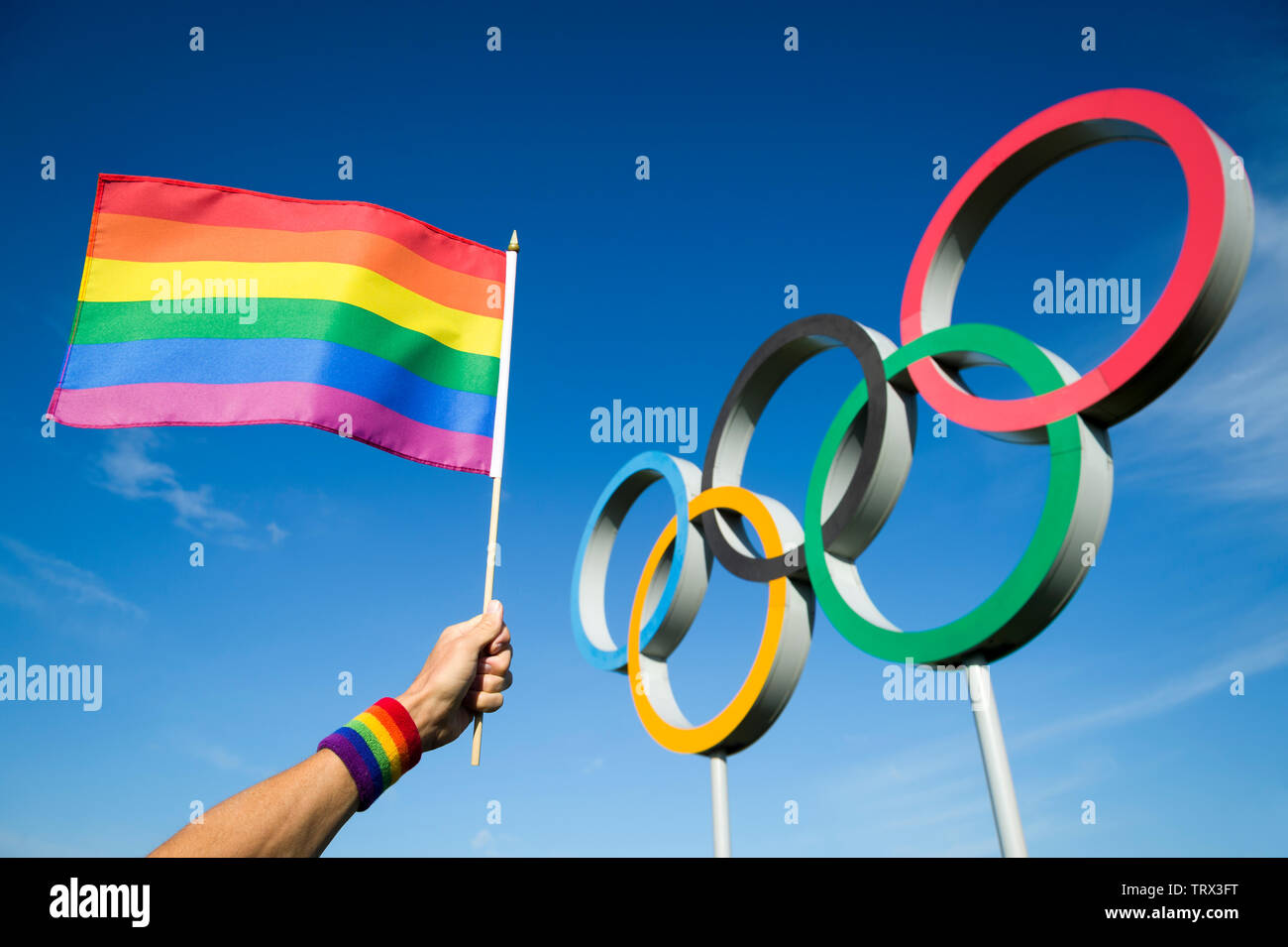 Londres - 4 mai 2019 : une main portant un bandeau de couleur arc-en-ciel vagues gay pride flag est en face d'Anneaux olympiques contre ciel bleu. Banque D'Images