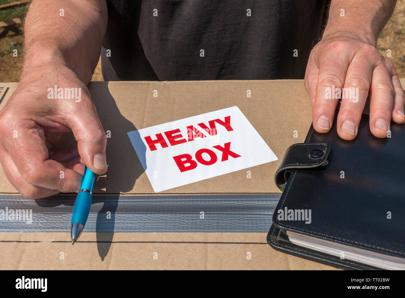 Les mains de l'homme offrant un stylo pour obtenir une signature pour la livraison d'une grande boîte en carton, avec un fort 'lourd' autocollant sur le dessus. Banque D'Images
