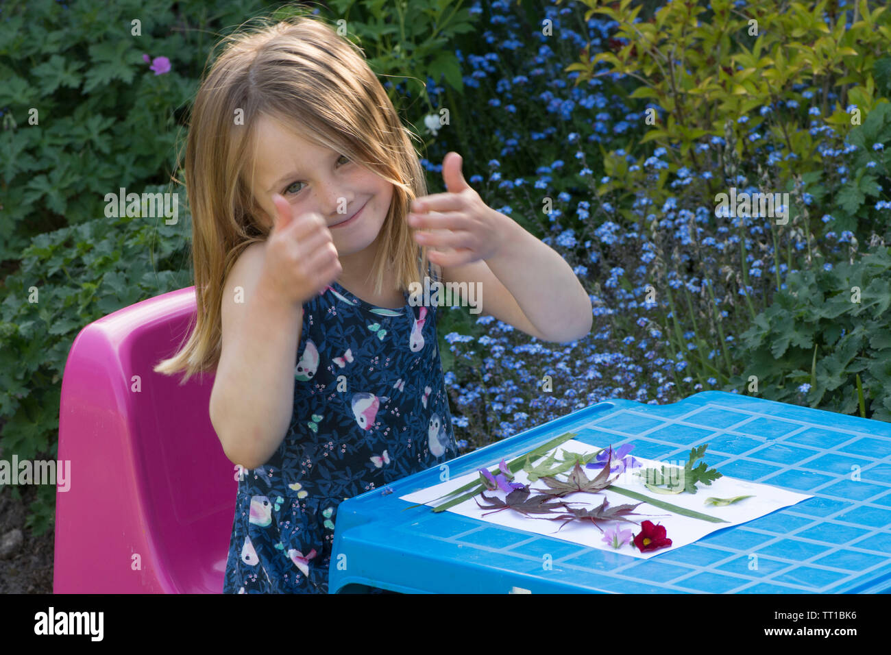 Trois ans, fille giving Thumbs up assis à une petite table, fier d'une photo faite à partir de fleurs et de feuilles de papier collées sur la nature, l'artisanat d'art Banque D'Images