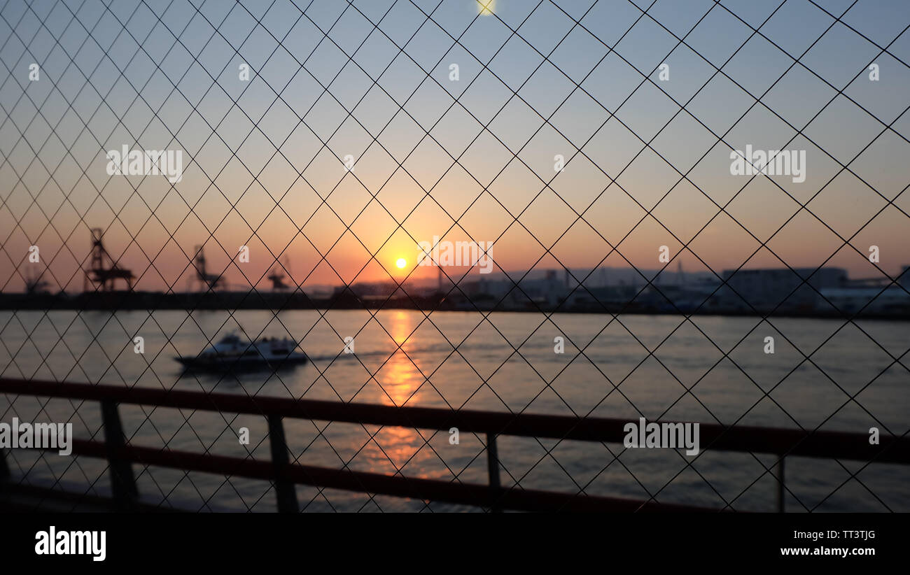 Grillage, avec arrière-plan flou de mer avec navire, contours de grues de chargement du port et les bâtiments, contre un coucher de soleil avec twilight dans le ciel, Banque D'Images