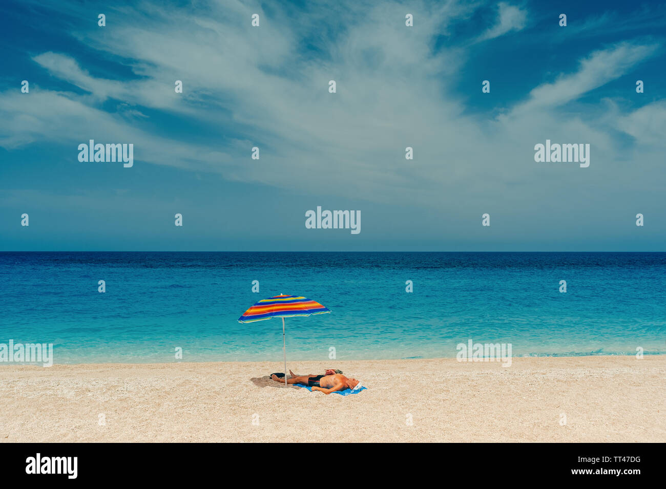 Vieil homme insouciant bronzer sur une plage de sable à l'ombre du parapluie à rayures avec jolie vue sur la mer. Banque D'Images