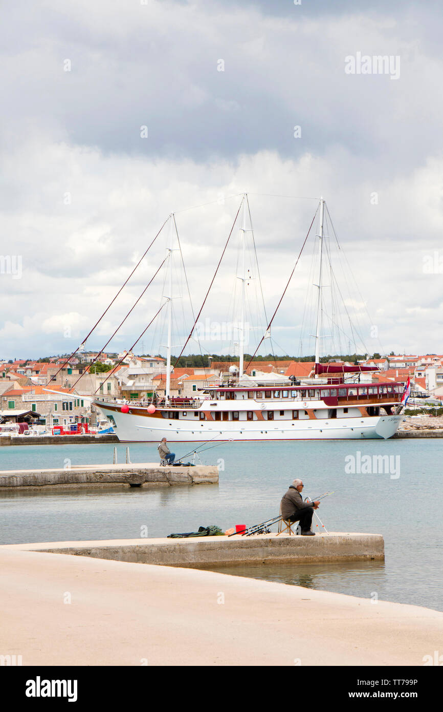 Vodice, Croatie - 30 Avril 2019 : deux hommes assis seul et patient sur la jetée en bord de pêche avec des cannes à pêche, et un voilier amarré derrière eux Banque D'Images