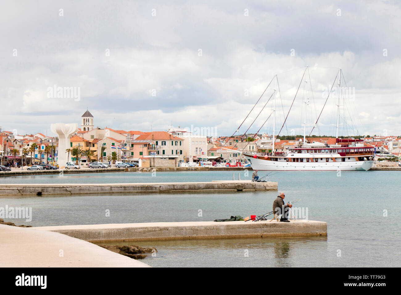 Vodice, Croatie - 30 Avril 2019 : deux hommes assis seul et patient sur la jetée en bord de pêche avec des cannes à pêche, et un voilier amarré derrière eux w Banque D'Images