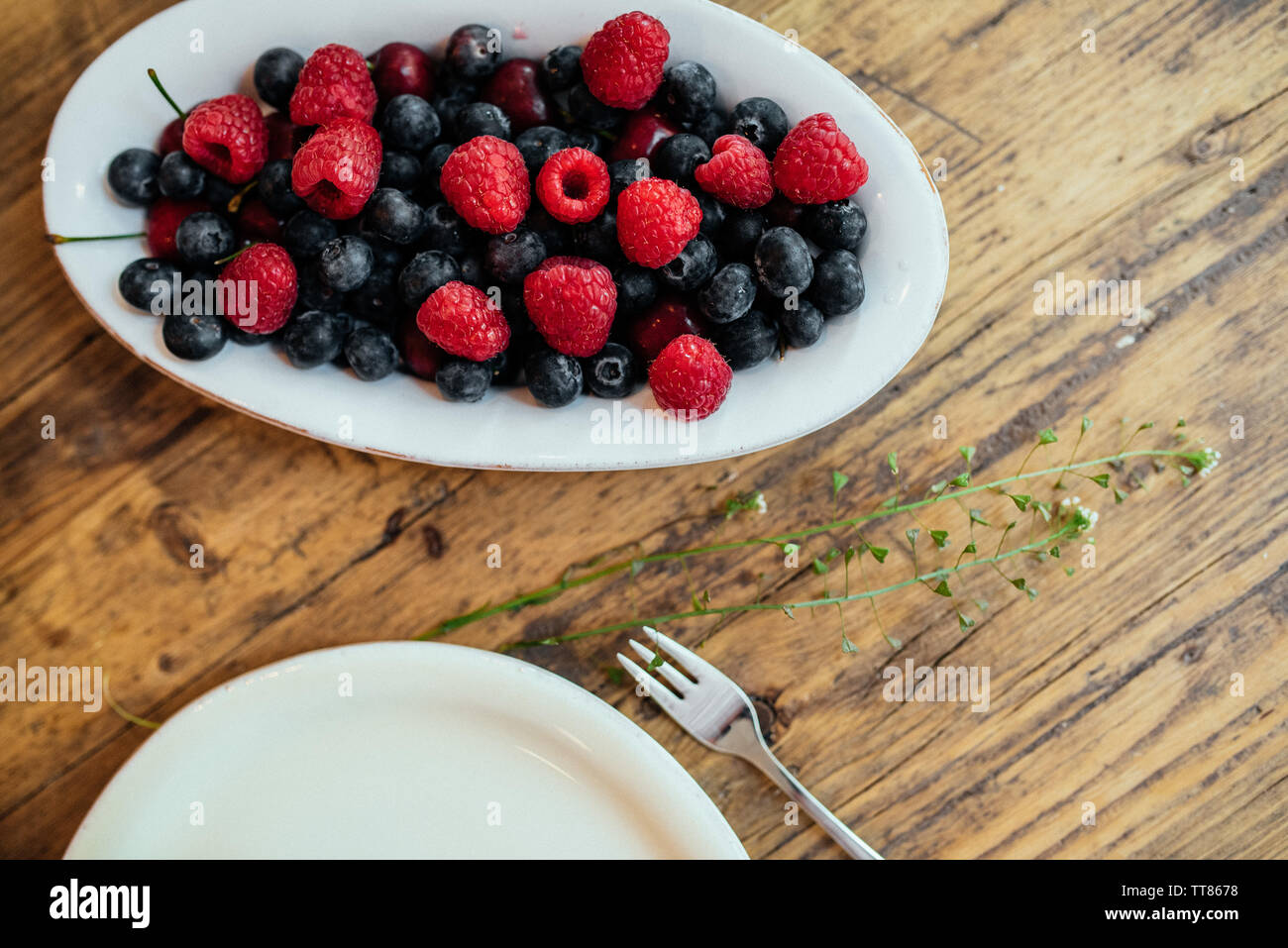 Les bleuets et les framboises dans une assiette blanche, décorée sur une table en bois avec des plantes et une fourchette Banque D'Images