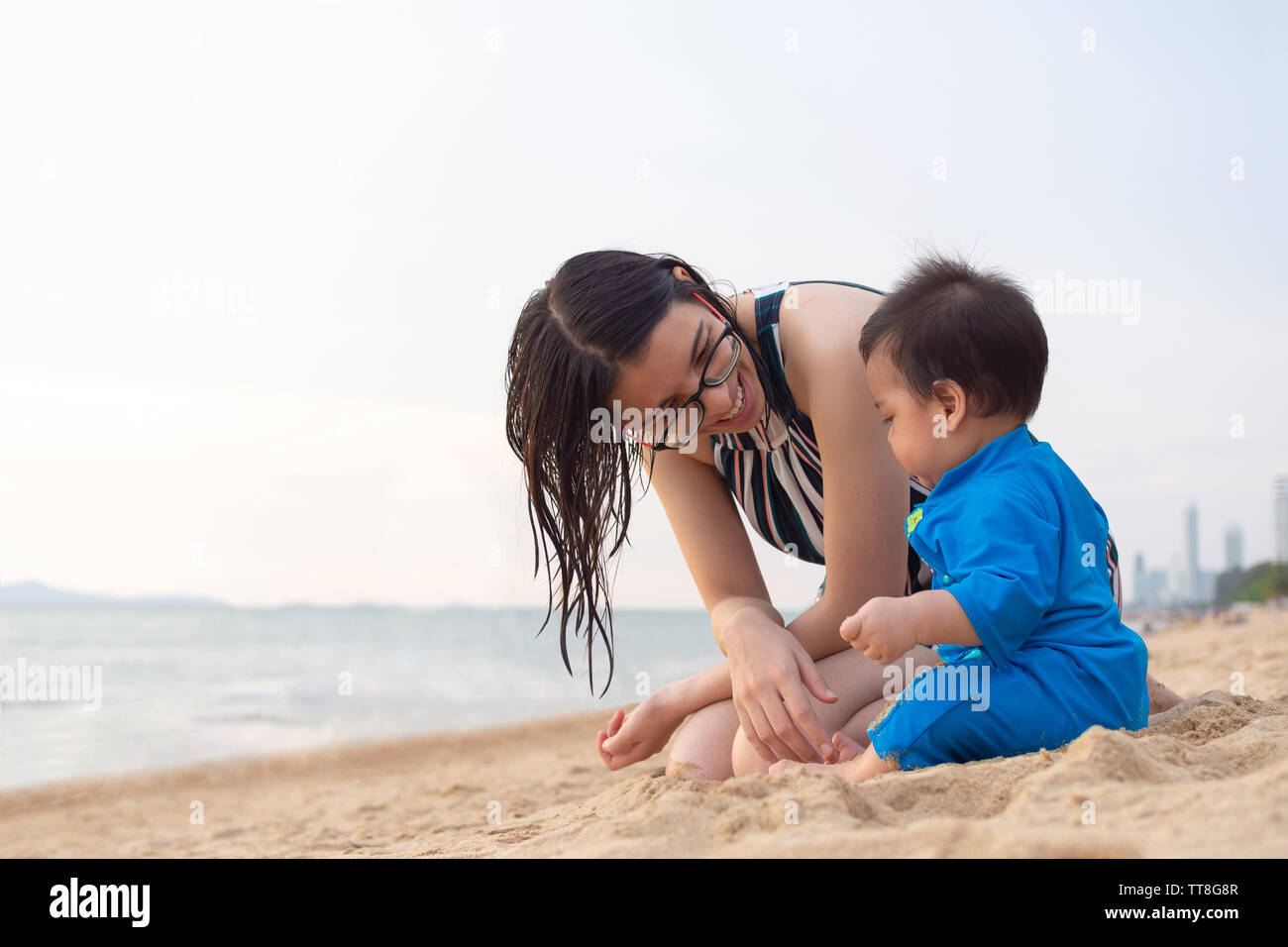 Heureux jeune mère souriant et regardant son petit bébé garçon jouant sur le sable de la plage tropicale à l'été. Les gens de vie familial et trave Banque D'Images