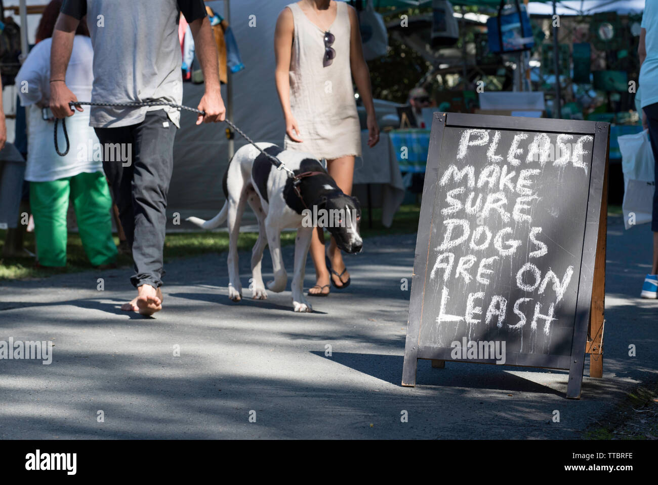 Personnes et un chien à l'agriculteur d'un week-end au marché communautaire Pacific Palms, Nouvelle Galles du Sud avec un signe lui demandant de garder leurs propriétaires de chien en laisse. Banque D'Images
