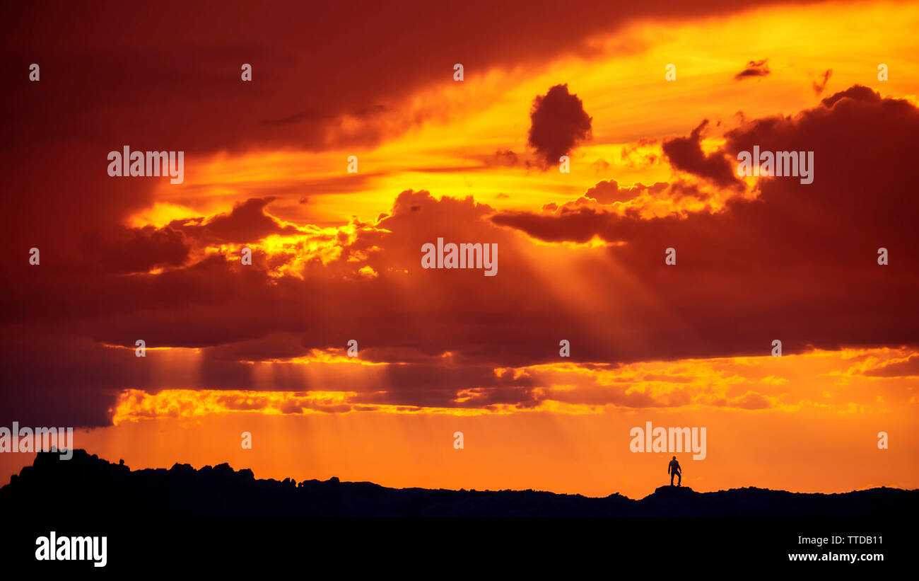 Silhouette Of Man Standing On Mountain Desert Ridge dans Arches National Park dans le cadre de spectaculaire coucher de soleil Nuages et rayons de soleil Banque D'Images