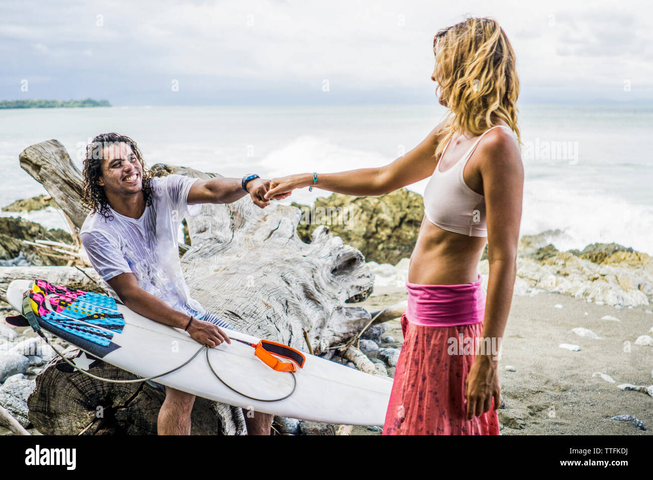 Heureux homme woman's hand holding surfer tout en restant assis sur driftwood at beach Banque D'Images