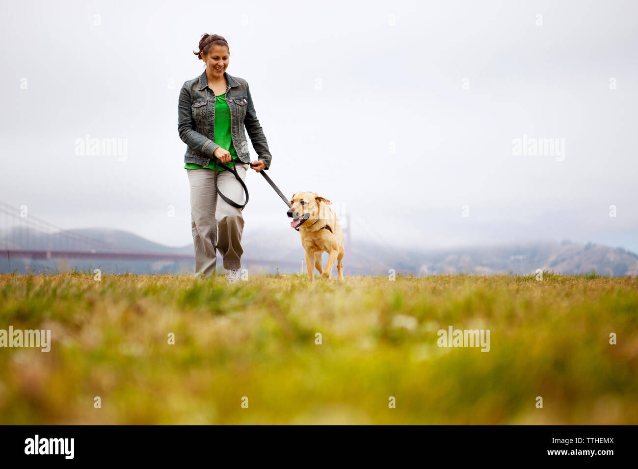 Happy woman walking with dog on grassy field against cloudy sky Banque D'Images