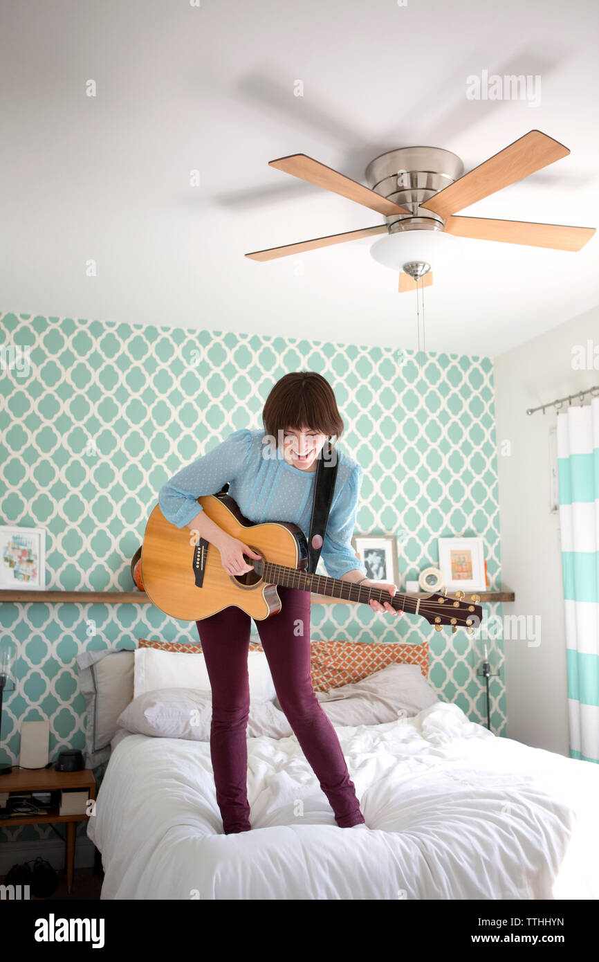 Happy woman playing guitar while standing on bed at home Banque D'Images