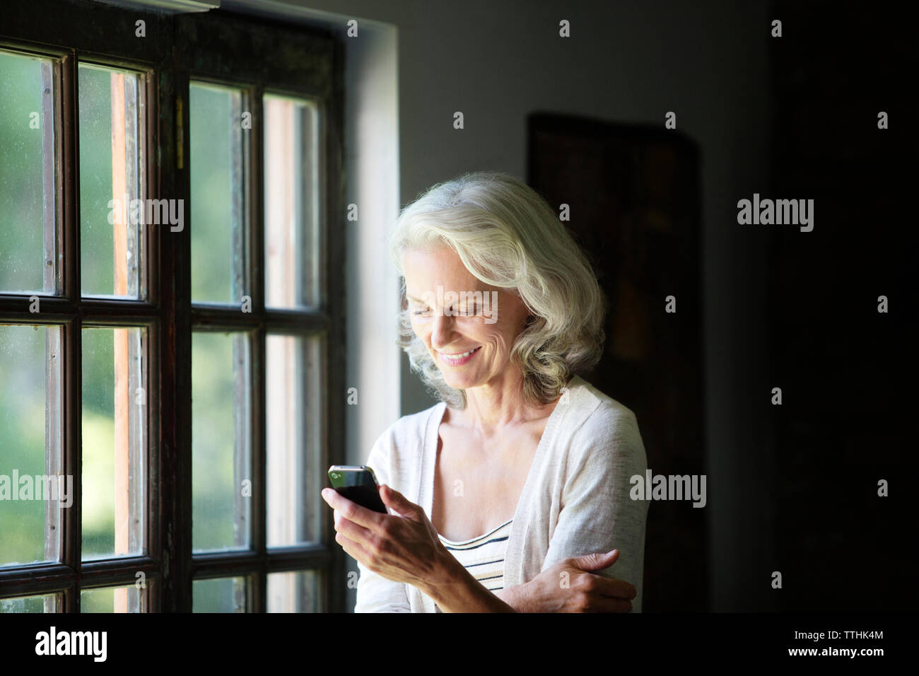 Happy senior woman using phone while standing par fenêtre à la maison Banque D'Images