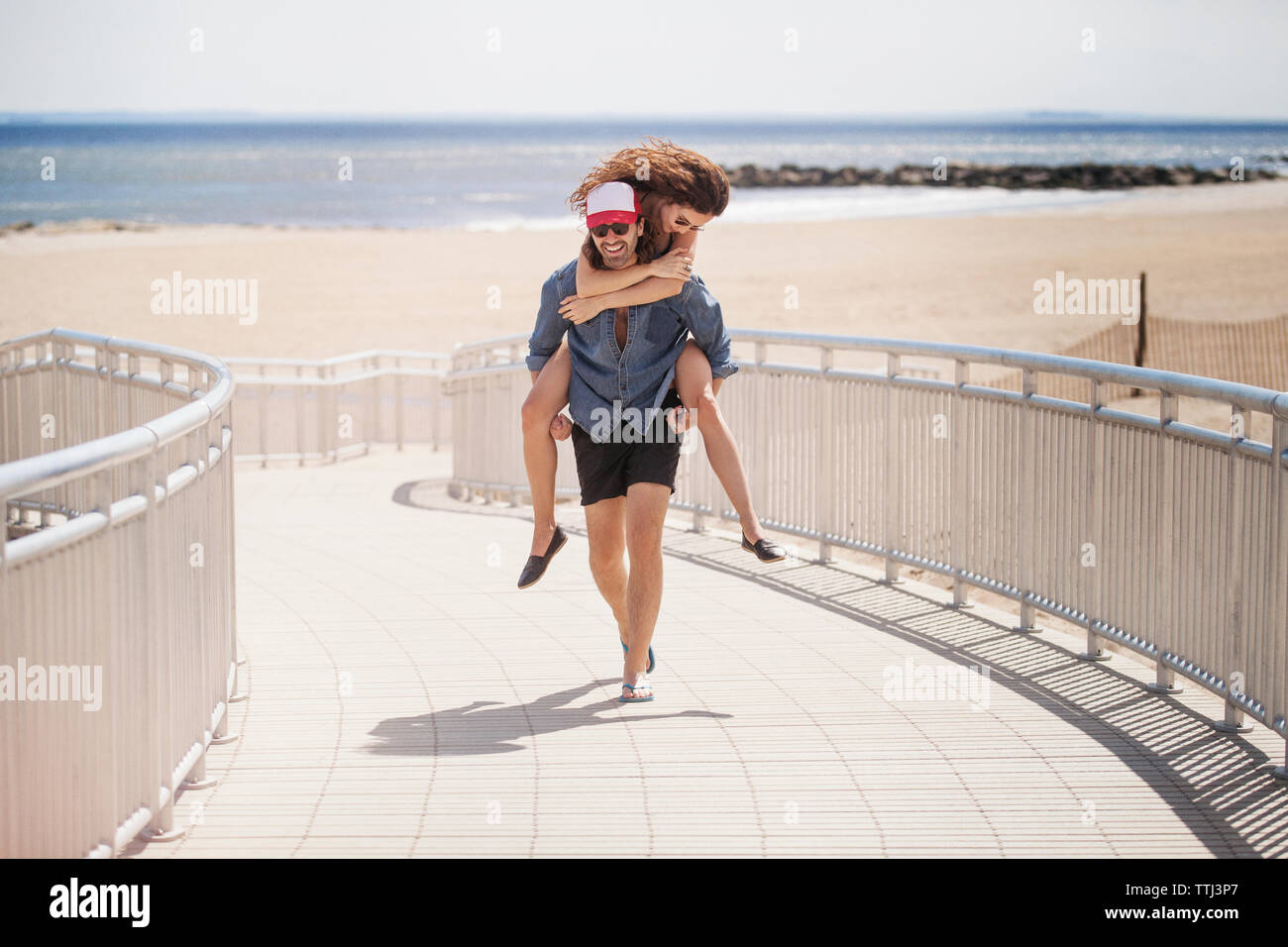 Happy man piggybacking woman en marchant sur boardwalk at beach Banque D'Images