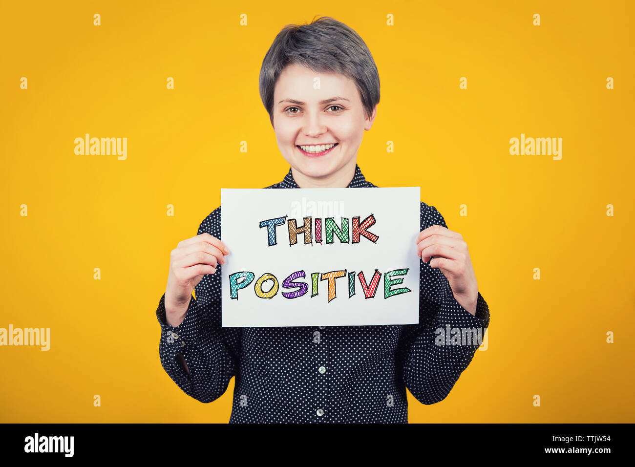 Extraverti optimiste hipster Jeune femme tenant une feuille de papier blanc avec le texte du message pensez positif plus isolé sur fond jaune. Mo colorés Banque D'Images