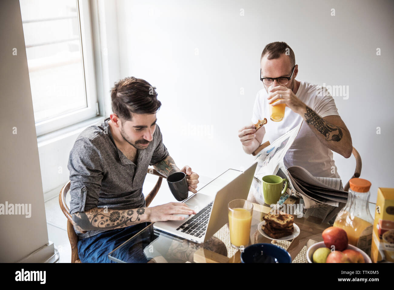 Les hommes gais à l'aide d'ordinateur portable et lisant le journal pendant le petit-déjeuner à la maison Banque D'Images