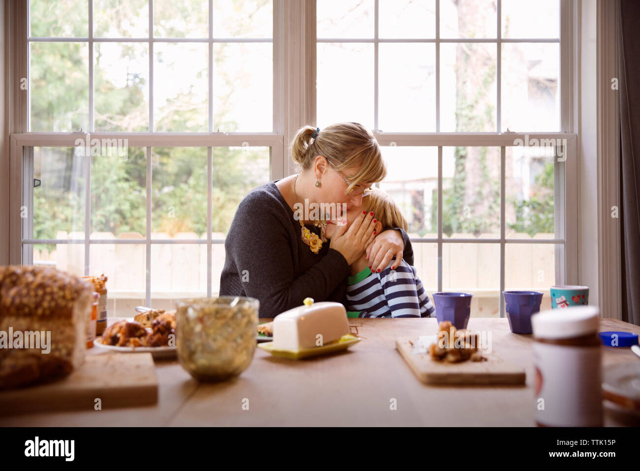 Mère embrassant fils assis à table des aliments contre la fenêtre dans salle de séjour Banque D'Images
