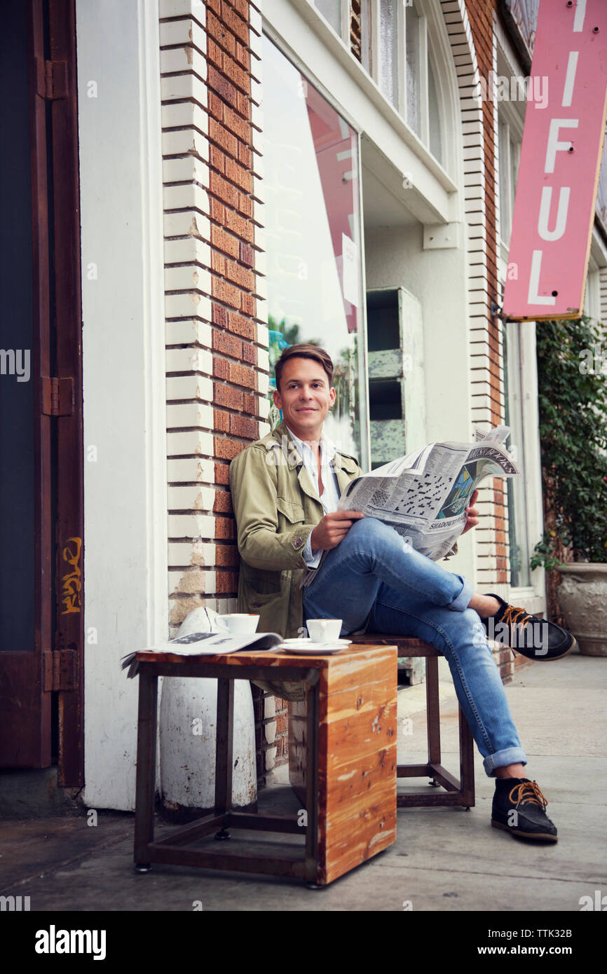 L'homme avec du papier journal à la route en position assise contre store Banque D'Images