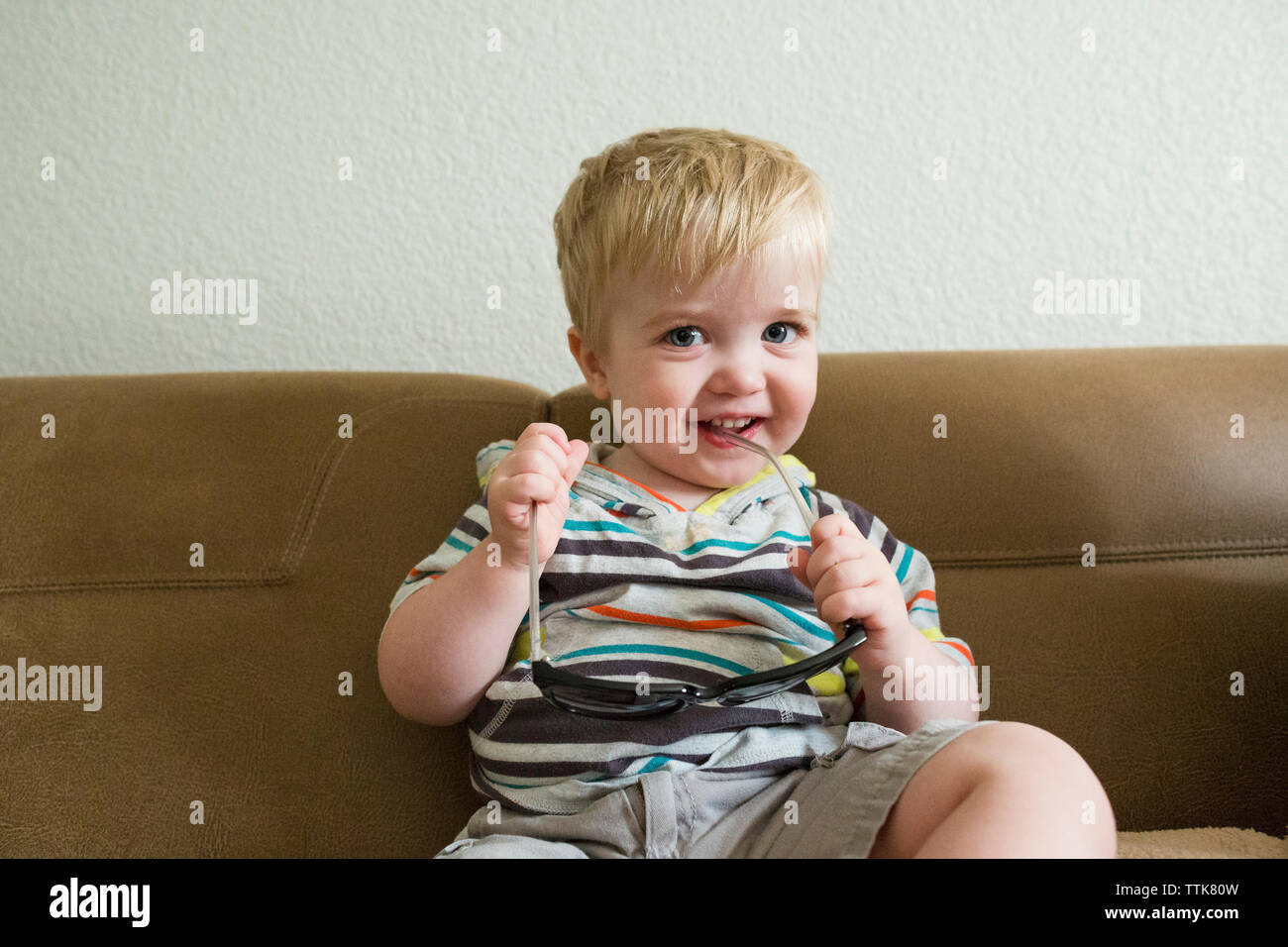 Portrait of cute happy baby boy holding sunglasses, assis sur un canapé contre le mur à la maison Banque D'Images