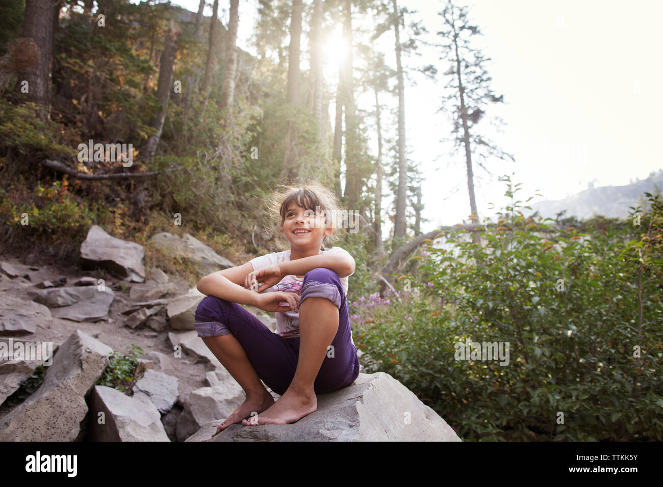 Happy girl sitting on rock against trees in forest Banque D'Images