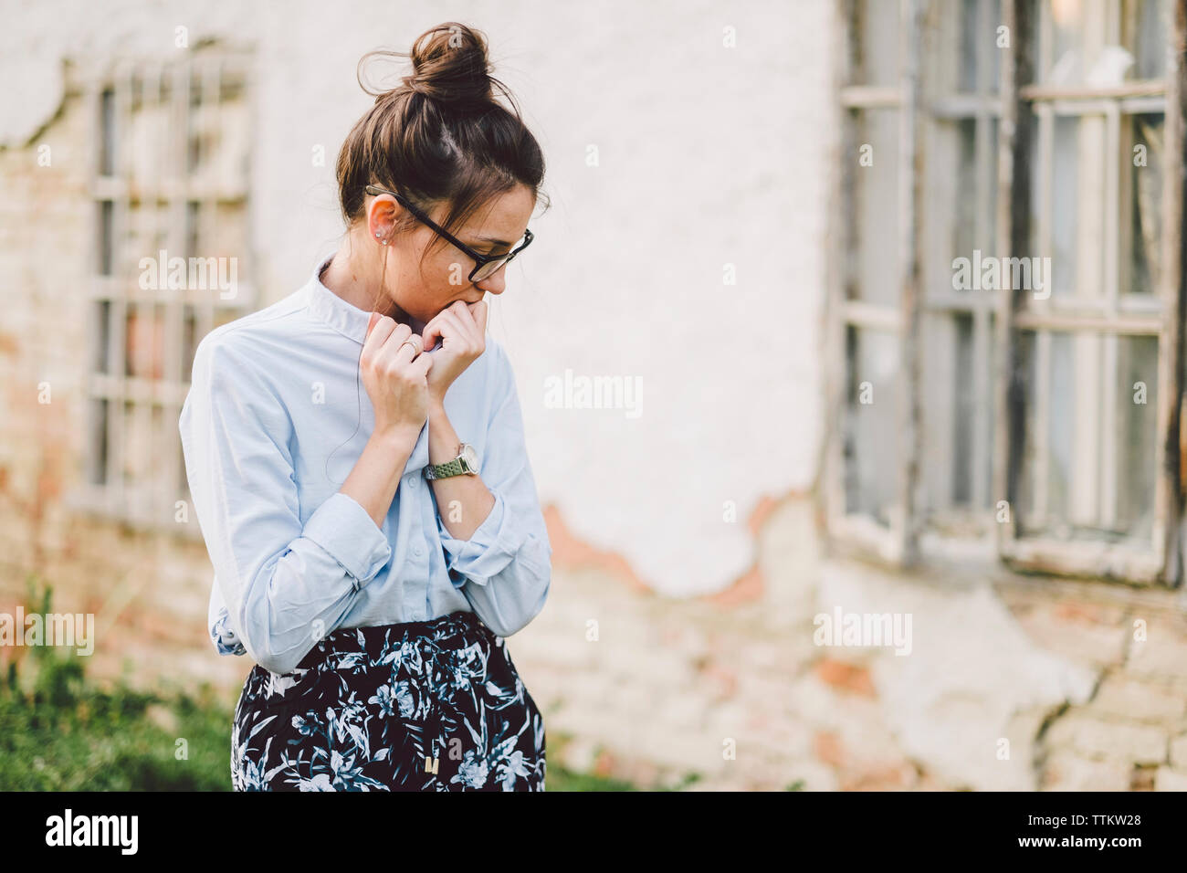 Sad young woman wearing eyeglasses standing by old weathered building at park Banque D'Images