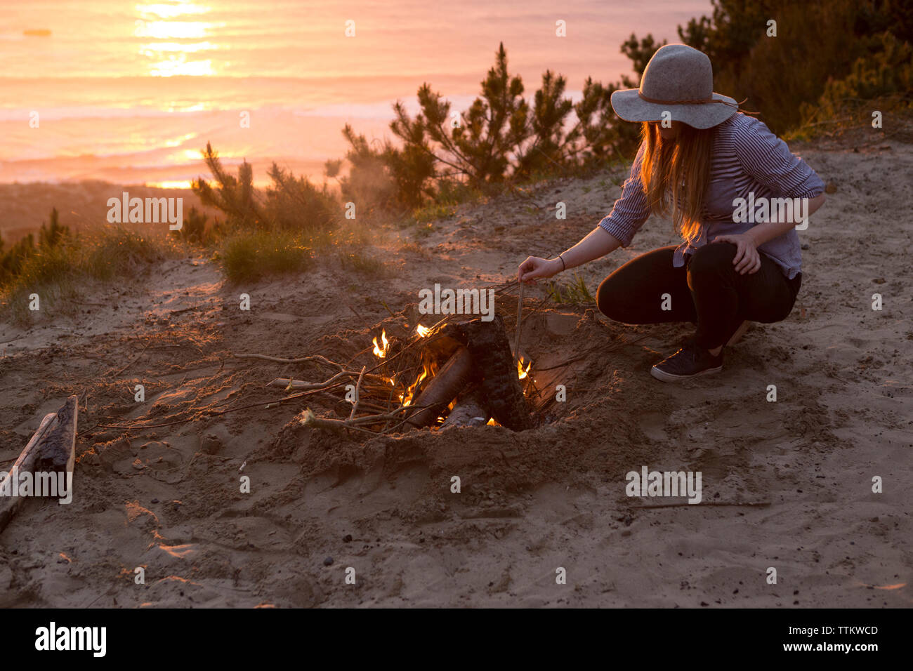 Woman crouching par feu de camp à la plage pendant le coucher du soleil Banque D'Images
