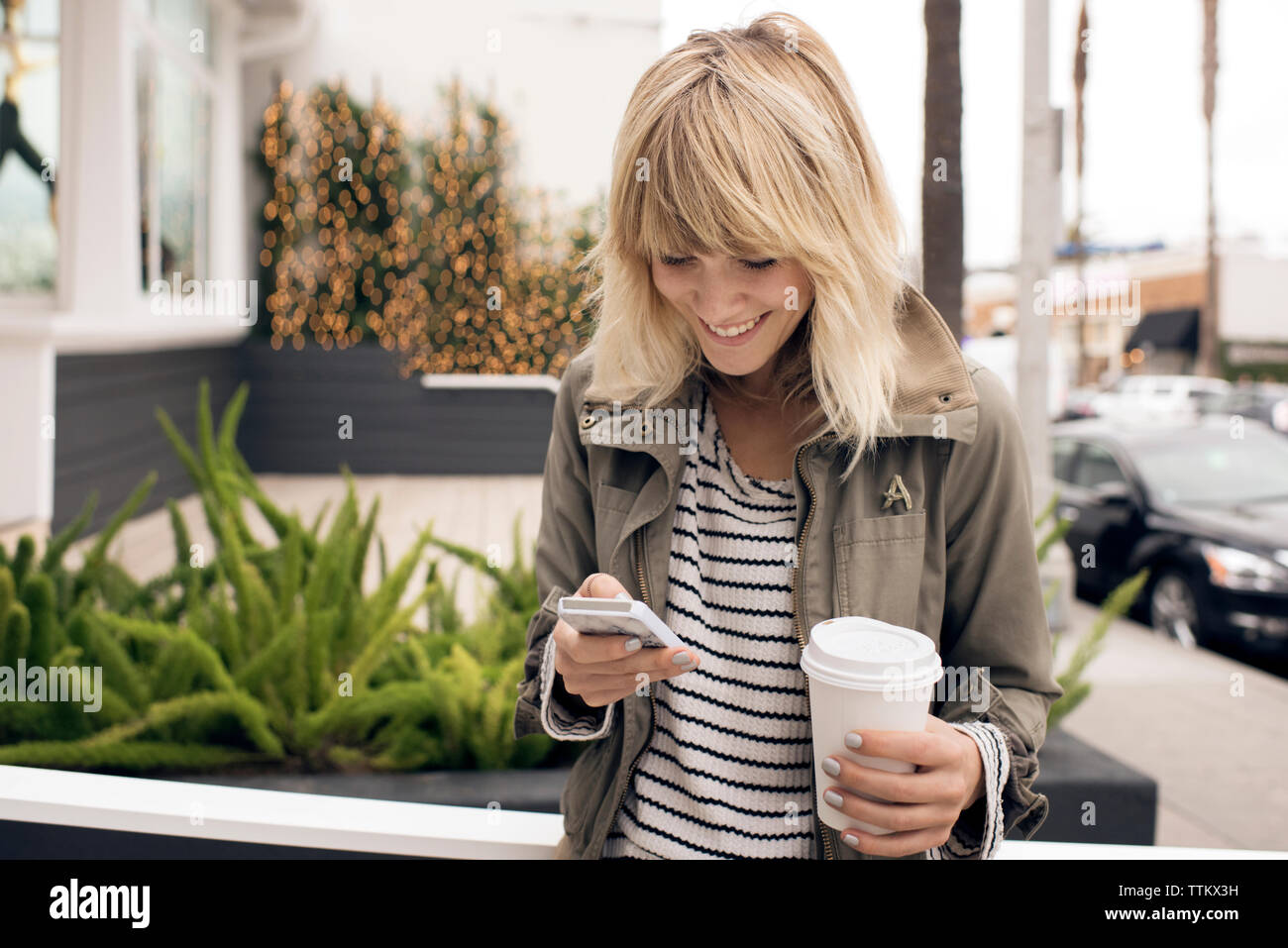 Happy young woman using smart phone en maintenant le verre jetable par city street Banque D'Images