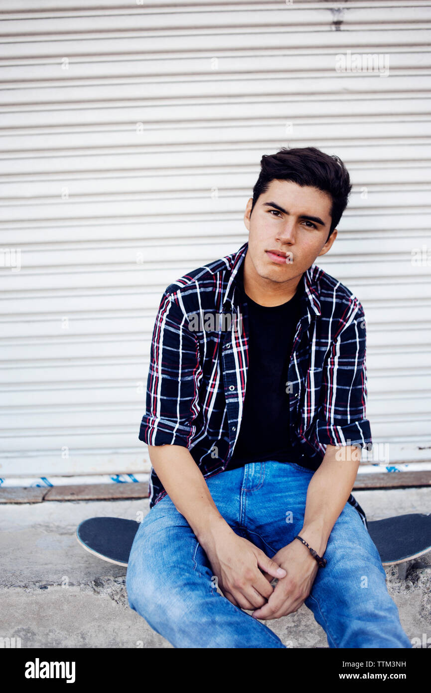 Portrait of smiling man sitting on skateboard contre l'obturateur fermé Banque D'Images