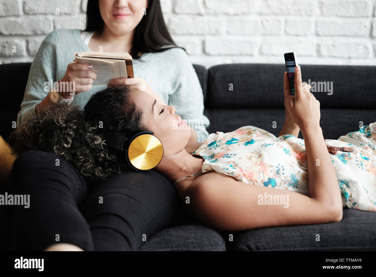 Femme à l'aide de téléphone mobile tout en vous relaxant sur les genoux de petite amie reading book in living room Banque D'Images