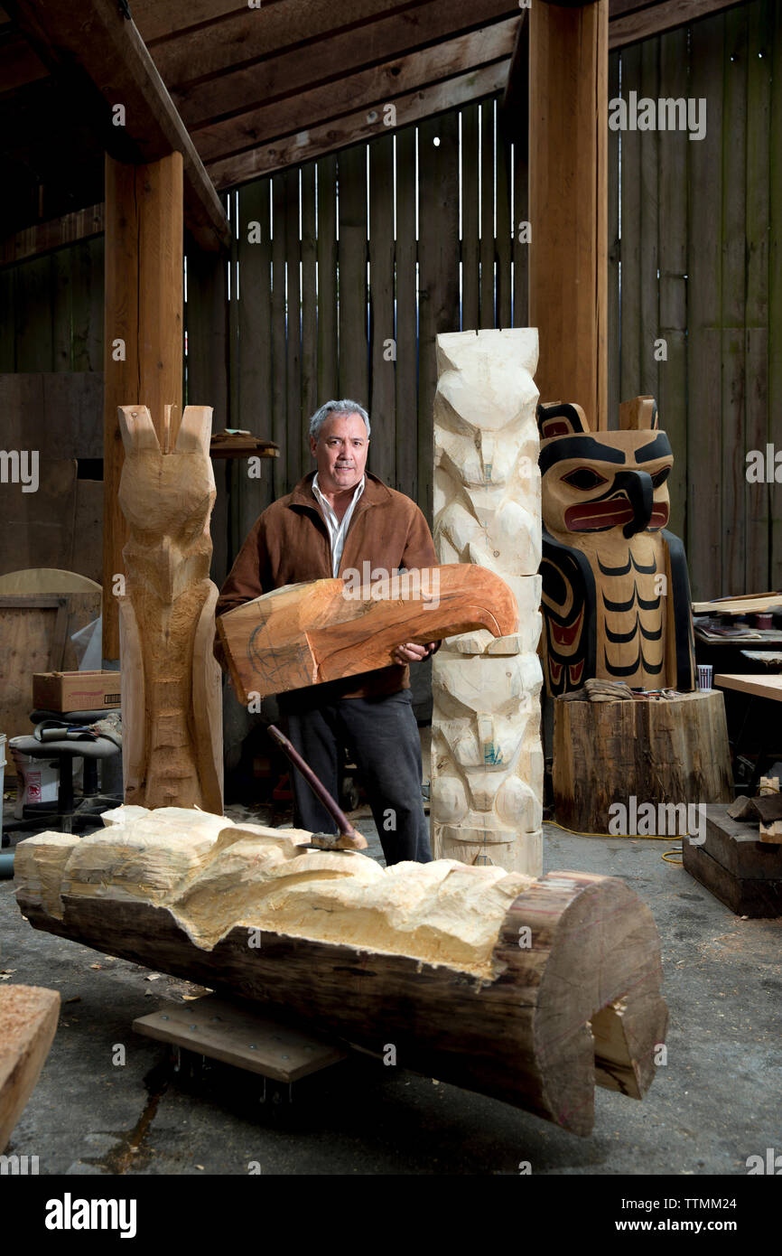 CANADA, Vancouver, Colombie-Britannique, le Haida Carver tient la tête d'un corbeau et se trouve à côté de totems à son magasin à Granville Island Banque D'Images