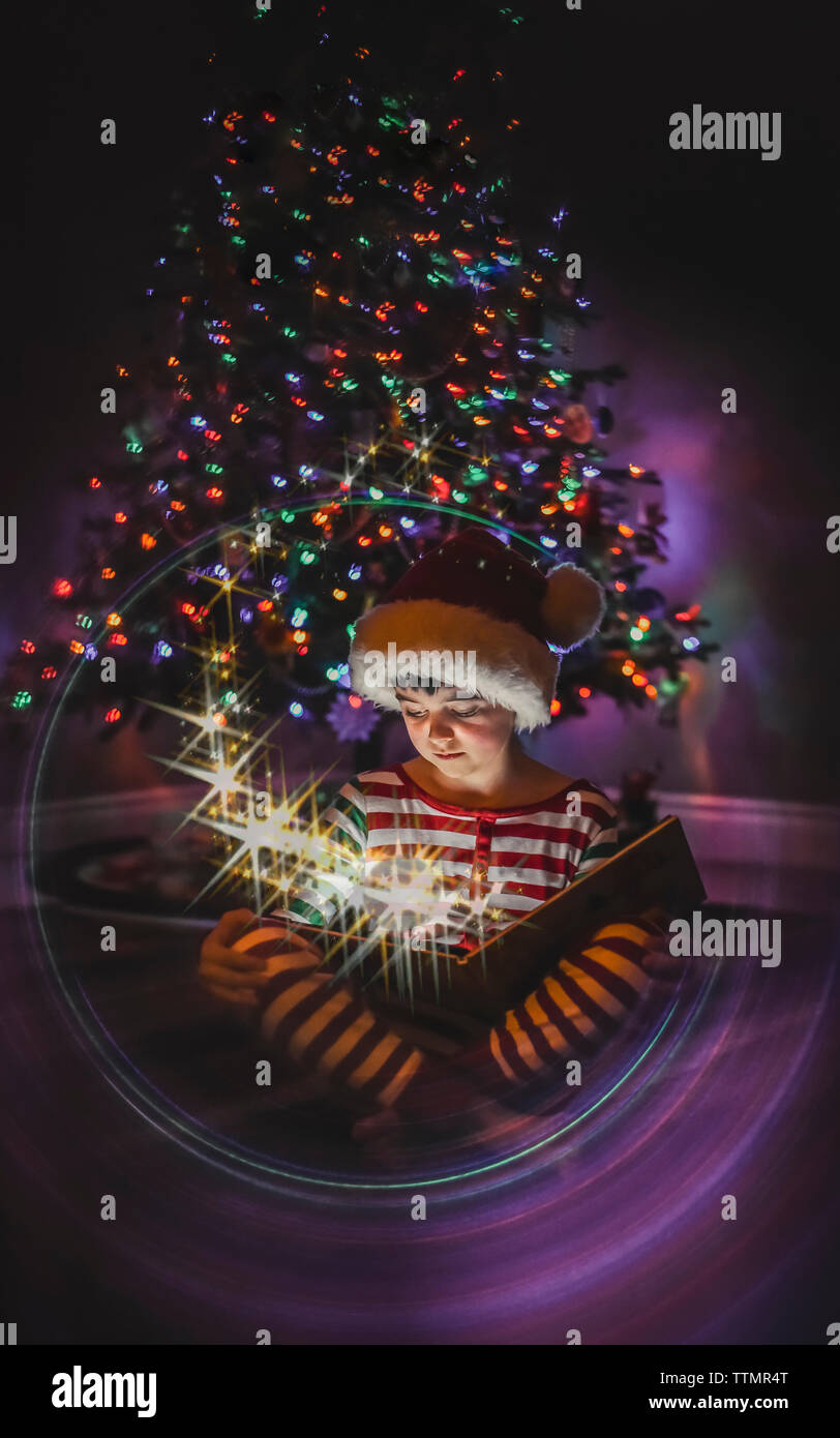 Boy reading livre magique en face de l'arbre de Noël avec visage illuminé. Banque D'Images