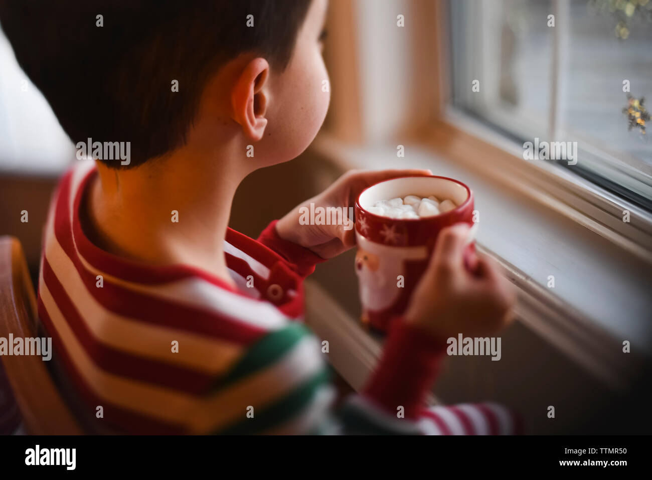 Jeune garçon en pyjama de Noël de boire un chocolat chaud en regardant par la fenêtre. Banque D'Images