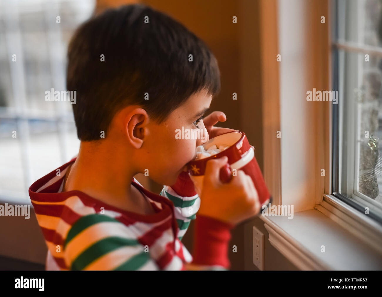 Jeune garçon en pyjama de Noël de boire un chocolat chaud en regardant par la fenêtre. Banque D'Images