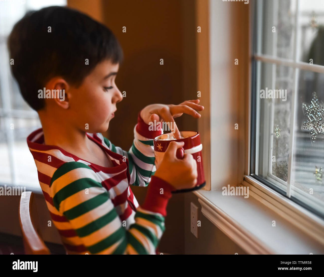 Boy holding tasse de chocolat chaud avec des guimauves assise à côté de fenêtre. Banque D'Images