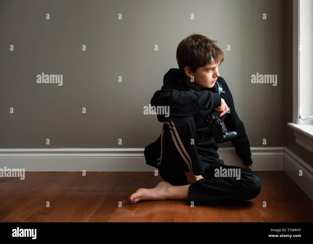 Boy sitting on floor looking out window avec contrôleur de jeu vidéo. Banque D'Images