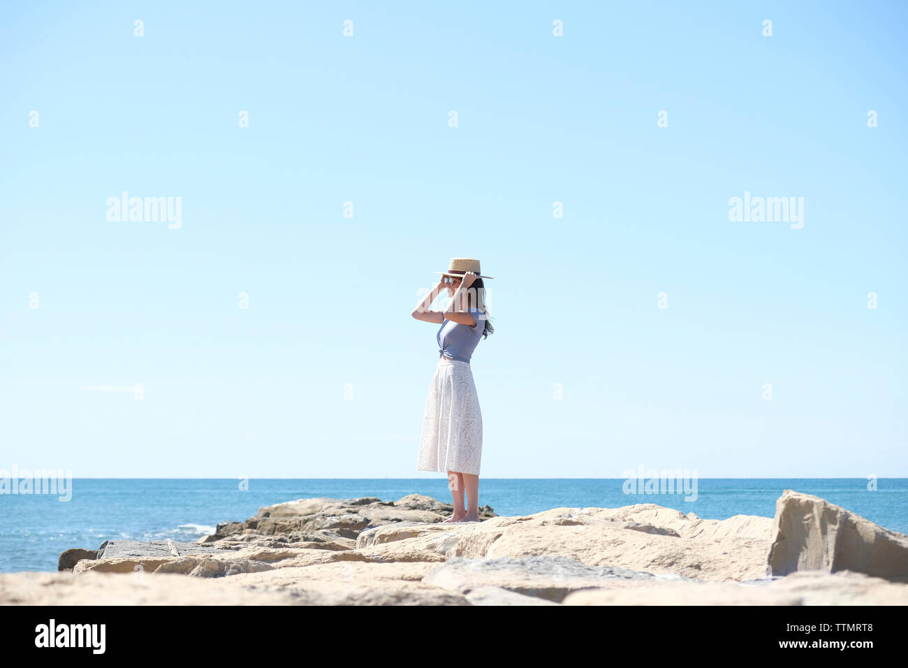 Young woman wearing hat en étant debout sur la plage de rochers à contre ciel clair au cours de journée ensoleillée Banque D'Images