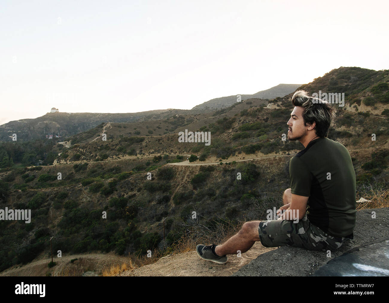 Toute la longueur du jeune homme à la vue au while sitting on rock contre le ciel au coucher du soleil Banque D'Images