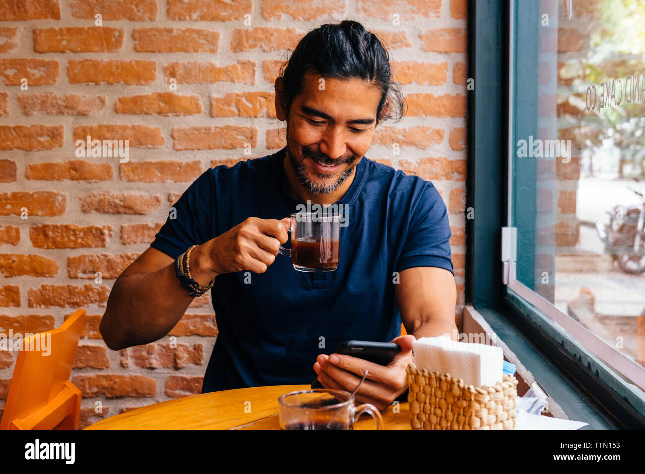 Smiling mature man avec du café noir à l'aide de smart phone against brick wall in cafe Banque D'Images
