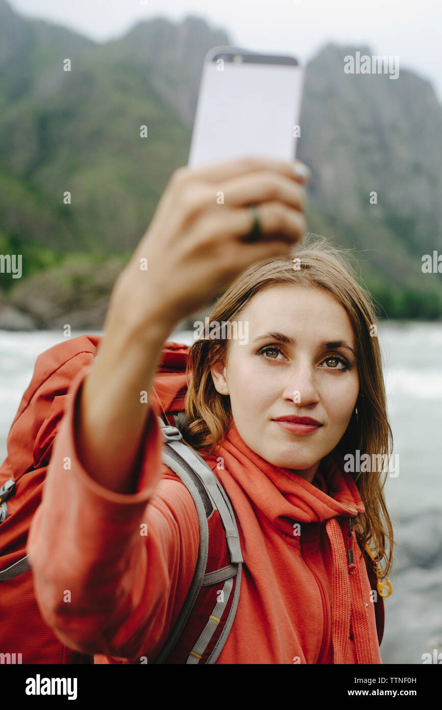 Female hiker en tenant avec selfies mobile phone while standing at riverbank contre mountain Banque D'Images