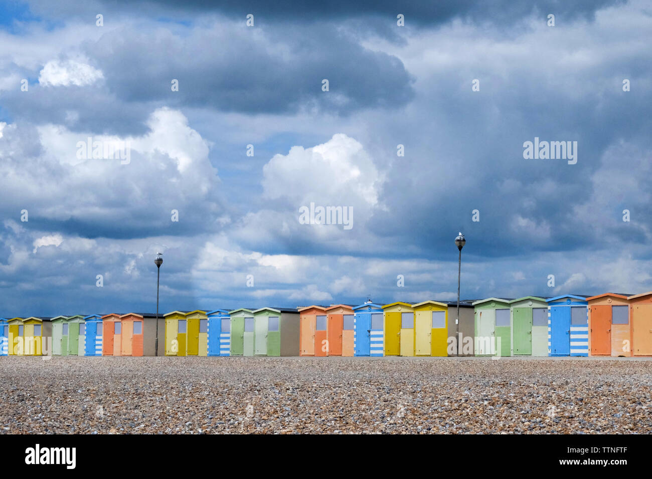 Une ligne de 22 cabines colorées qui traversent le centre de l'image ci-dessous, est une plage de galets et de jaune au-dessus est un formidable nuageux bleu et blanc Banque D'Images