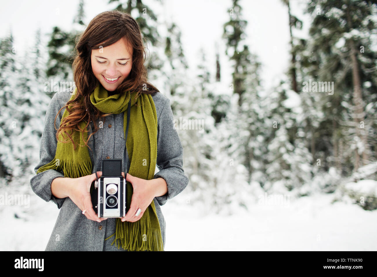 Smiling woman holding camera debout contre la neige a couvert de pins en forêt Banque D'Images