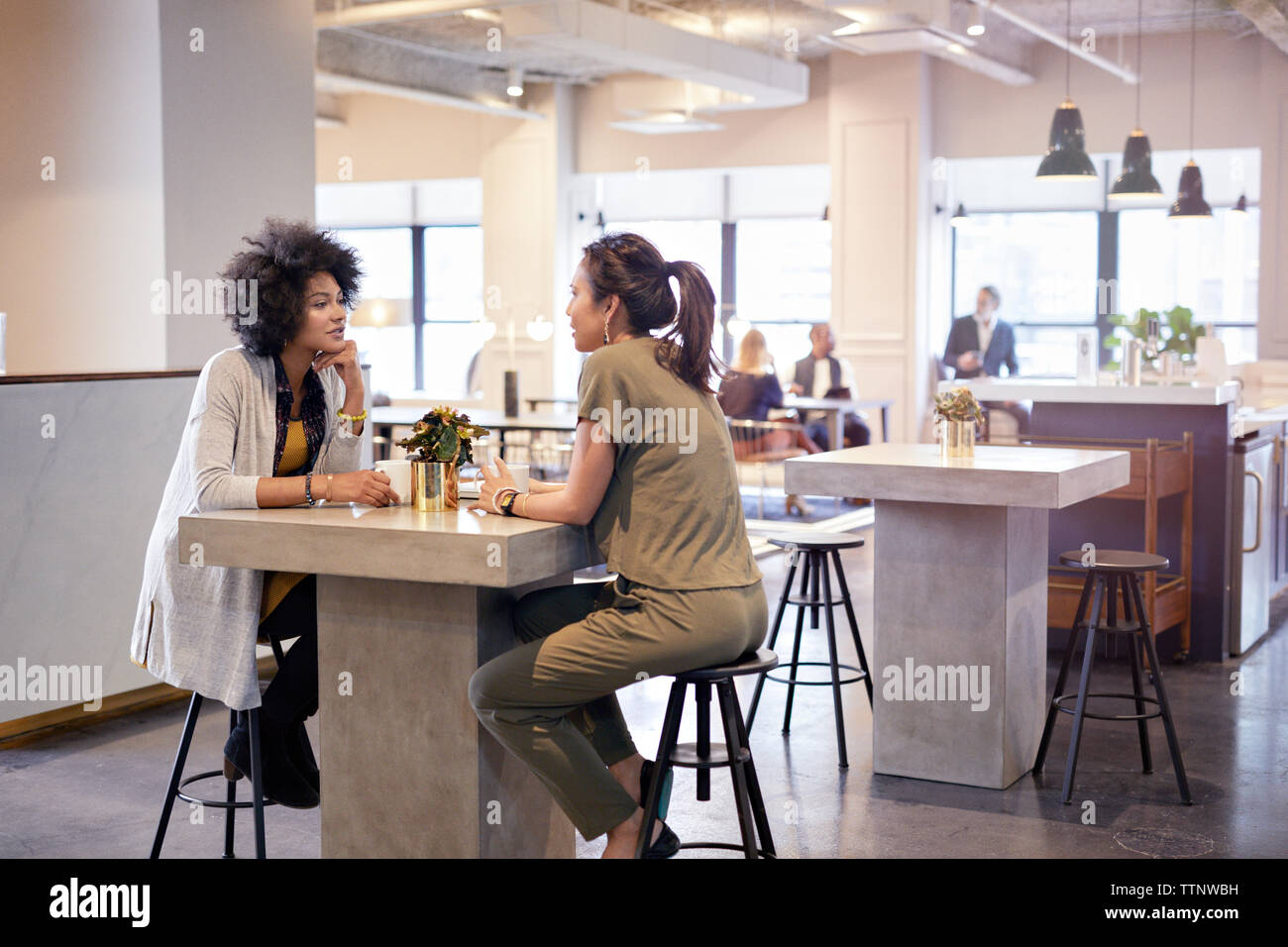 femmes d'affaires parlant tout en étant assis dans la cafétéria au bureau créatif avec des collègues en arrière-plan Banque D'Images